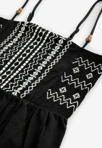 Svart topp med intrikata vita broderier i geometriska mönster. Justerbara band med trädetaljer. Åtsittande textur vid fållen.