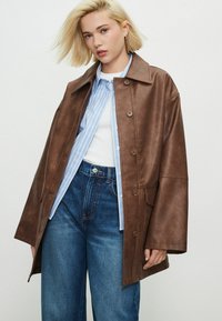 Jeune femme aux cheveux blonds courts portant une veste en cuir marron, une chemise rayée bleue, un haut blanc et un jean bleu taille haute sur un fond uni.