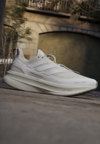 Scarpa da corsa Adidas bianca con tre strisce, suola spessa e imbottita etichettata Light Boost, mostrata all'aperto su pavimentazione in pietra.
