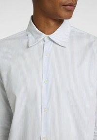Camisa blanca a rayas con líneas verticales azul claro, cuello abotonado y botones en la parte delantera, con una textura de tela suave.