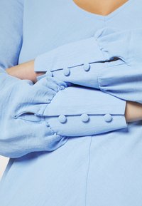 Blusa azul clara com decote em V e mangas longas bufantes, detalhada com botões discretos e um tecido suave e texturizado.