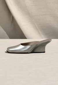 Chaussure mule métallique argentée avec un bout carré fermé et un talon compensé incurvé sur une surface beige clair.
