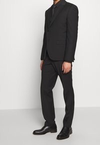 Hombre de pie vestido con un traje negro ajustado, camisa negra, corbata negra y zapatos de vestir de cuero negro, contra un fondo claro liso.