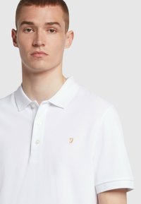 Witte polo shirt gemaakt van gestructureerde stof, met een drieknopenlijst en een klein gouden geborduurd logo op de linkerborst.