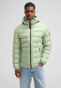 Giacca a vento verde chiaro con cappuccio, dotata di zip frontale, dettagli neri e due tasche laterali. Design trapuntato orizzontale.