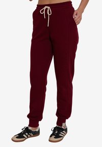 Burgundowe dresy z elastycznym pasem, kieszeniami po bokach i zwężanymi nogawkami, wykonane z miękkiego materiału. Połączone z czarno-beżowymi trampkami.