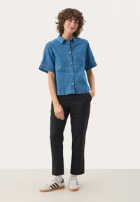 Camicia di jeans a maniche corte di colore azzurro chiaro con taschino, chiusura con bottoni e dettagli sulle cuciture, abbinata a pantaloni neri e sneakers bianche.