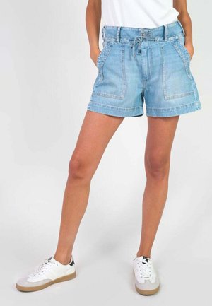 Kvinde iført lyseblå denimshorts med forlommer, hvide sneakers og en hvid top, stående med hænderne i lommerne.
