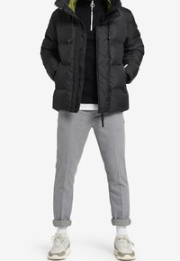 Musta toppatakki, jossa on vihreä vuori, vetoketjullinen etuosa ja kiristysnyörillinen huppu, yhdistetty vaaleanharmaisiin housuihin ja kermaan vivahtaviin tennareihin.