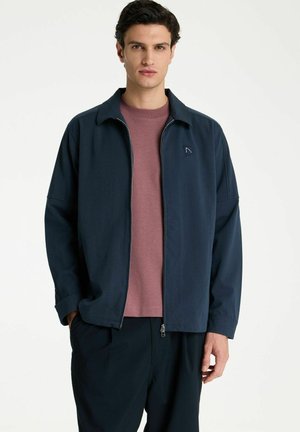 Jonge man draagt een marineblauw jasje met rits over een mauve t-shirt en donkere broek, staand met handen in de zakken tegen een neutrale achtergrond.