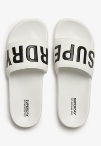 Hvide slidesandaler med en tyk sort rem, der viser ordet "SUPERDRY." Glat finish, flad sål og 50% genanvendte materialer.