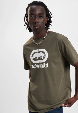 Young man with dreadlocks wearing an olive green Ecko unltd. t-shirt and silver chain necklace, standing against a plain white background.
