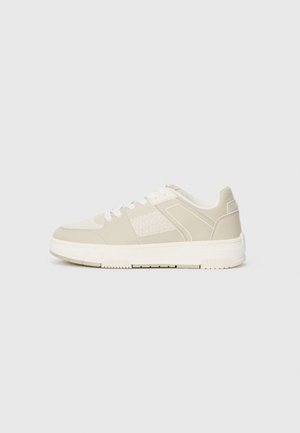 Beige sneakers with textured overlays, white laces, and a thick, textured white sole. Features geometric design accents on sides.