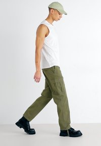 Pantaloni cargo verde oliva con dettagli in zip, abbinati a una canotta bianca aderente e stivali neri chunky, indossati con un cappellino verde chiaro.