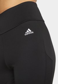 Svarta leggings i slät, elastisk tyg med en bred midjeresår och en liten vit Adidas-logotyp på övre sidan. Sidopaneldetaljer synliga.