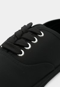 Svart sneaker med tykke svarte snører trådet gjennom fire sølvfargede øyeletter på en enkel bakgrunn.