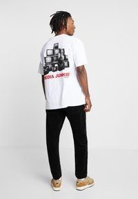 T-shirt en coton blanc avec un graphisme noir et blanc de télévisions empilées au dos et "MEDIA JUNKIE" en texte rouge en dessous.