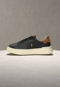 Baskets basses noires avec semelle crème, languette de talon marron et petit logo brodé sur le côté, présentées sur une surface claire.