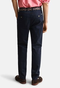 Hombre con pantalones azul marino oscuro, cinturón marrón tejido, camisa de manga larga rosa remangada y zapatos de ante marrón oscuro, de espaldas.