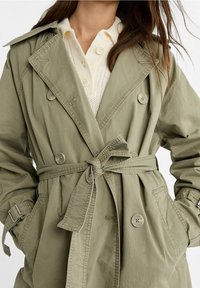 Olivinvihreä, vyöllä varustettu trench coat, jossa on suuret napit, hartiasuojat ja etutaskut. Valmistettu kevyestä kankaasta ja rento istuvuus.