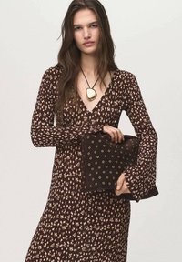 Robe florale marron à manches longues, décolleté en V profond et corsage ajusté. Sac à main noir avec des accents en œillets dorés. Collier en pendentif circulaire doré.