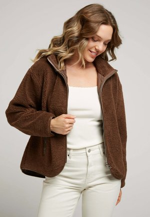 Veste zippée en polaire marron avec un col haut, doublure intérieure contrastante et coupe décontractée, associée à un haut blanc et à un pantalon de couleur claire.