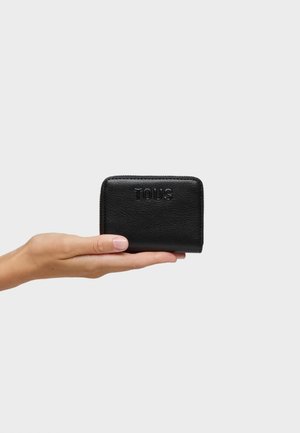 Mano che tiene un portafoglio nero in pelle con cerniera, impresso con il nome del marchio "TOUS", su uno sfondo grigio chiaro.