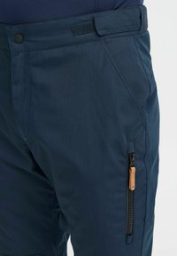 Pantaloni bleumarin dintr-un material ușor, cu un buzunar lateral cu fermoar și o bandă de piele pentru tragere, precum și o talie plată cu închidere Velcro.