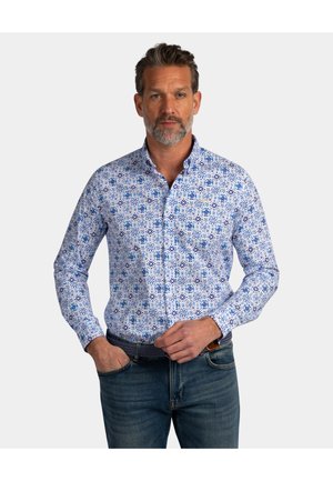 Middle-aged man with salt-and-pepper beard wearing a blue patterned button-up shirt and jeans, standing with one hand in pocket.