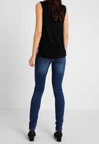 Mujer vistiendo jeans ajustados azul oscuro, top sin mangas negro y zapatos de tacón negros, de pie de espaldas contra un fondo liso.