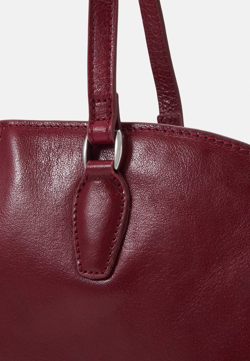 Gros plan d'une bandoulière de sac à main en cuir rouge foncé attachée avec un anneau métal argenté et une couture détaillée sur une surface en cuir lisse.