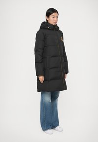 Melns pūkajams mētelis ar kapuci, rotāts ar zelta rāvējslēdzēju un logotipu. Mētelim ir kviltēts dizains, un tas ir kombinēts ar gaiši zilām plata kāju džinsiem.