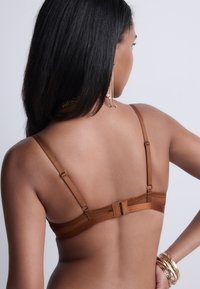 Lingerie marron avec des bretelles réglables et une fermeture à crochets. Texture lisse avec des détails de couture minimaux. Le dos du modèle est visible.