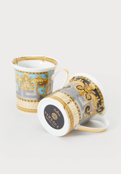 Porzellantassen zeigen filigrane goldene und blaue Muster mit barocken Akzenten. Eine davon trägt ein schwarzes Etikett mit der Aufschrift "VERSACE" und bunten Designs.