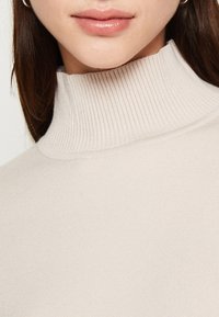 Pull à col roulé côtelé beige avec une coupe ajustée, présentant une texture verticale et un col haut qui se replie.