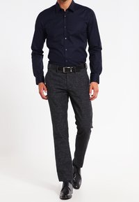 Camisa de botones azul marino de manga larga, combinada con pantalones oscuros de textura gris y zapatos de vestir negros, con un diseño ajustado y tela suave.
