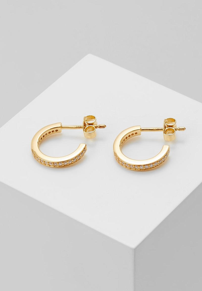 Paire de petites boucles d'oreilles créoles en or avec des pierres transparentes incrustées, présentées sur un cube blanc avec un fond gris.