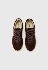 Ζευγάρι σκουρόχρωμων καφέ σουέντ αθλητικών παπουτσιών με χονδρές κορδόνια ίδιου χρώματος και μπεζ σόλες, φαίνονται από πάνω σε ανοιχτό φόντο.