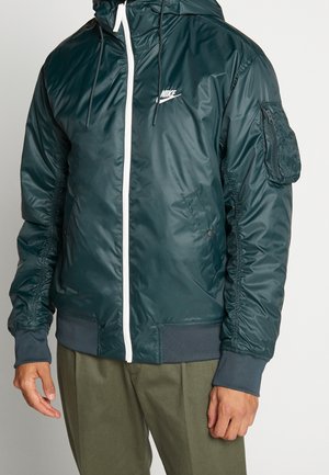 Mężczyzna w ciemnozielonej, zapinanej na zamek kurtce z kapturem marki Nike z zamykaną kieszenią na lewym rękawie oraz w oliwkowych spodniach.