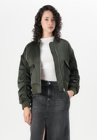 Maslinasto zelena bomber jakna, skraćeni dizajn, sjajna saten tekstura, s patentnim zatvaračem, rebrastim manžetama i bočnim džepovima. Uparena s sivom suknjom.