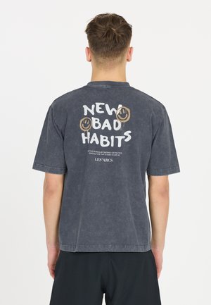 Grå grafisk t-shirt med korte ærmer. Har teksten "NEW BAD HABITS" og smilende ansigt på ryggen. Lavet af blødt, tekstureret stof.