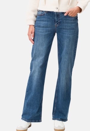 Person wearing blue straight-leg jeans, white sneakers, and a cream cardigan with one hand in pocket against a plain background.