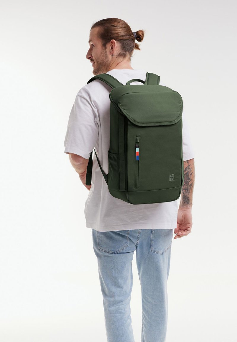Sac à dos en tissu vert avec un design rectangulaire, des bretelles ajustables, une poche zippée à l'avant et une étiquette d'accent colorée.