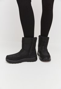 Botas negras impermeables con una cremallera en el lateral, parte superior acolchada y suela texturizada. Presentan una punta redondeada y una altura hasta la mitad de la pantorrilla.