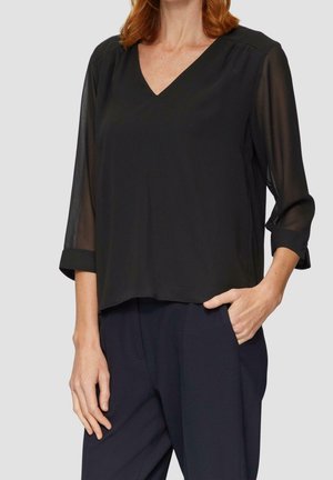 Femme portant un chemisier noir à col en V avec des manches trois-quarts transparentes et un pantalon foncé, une main dans la poche et l'autre détendue sur le côté.