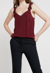 Mujer vestida con una blusa sin mangas de color burdeos con grandes detalles de anillos negros en las tirantes y pantalones negros, de pie con las manos en las caderas.