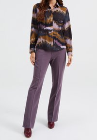 Långärmad skjorta med knappar i ett flerfärgat zigzag-mönster. I kombination med figurnära lila byxor. Modellen bär vinröda skor.