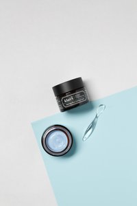 Svart krukke med Klairs Midnight Blue Calming Cream og en lys blå krem inni, sammen med en klar applikator, plassert på et blått ark.