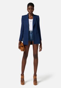 Blazer imbottito blu con spalle strutturate, abbinato a pantaloni di denim e scarpe con zeppa marroni. Tiene una pochette marrone.