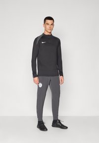 Svart långärmad träningsshirt med halvmatch och grå accenter, i kombination med grå byxor som har en logotyp, allt tillverkat av lätt, andningsbart material.